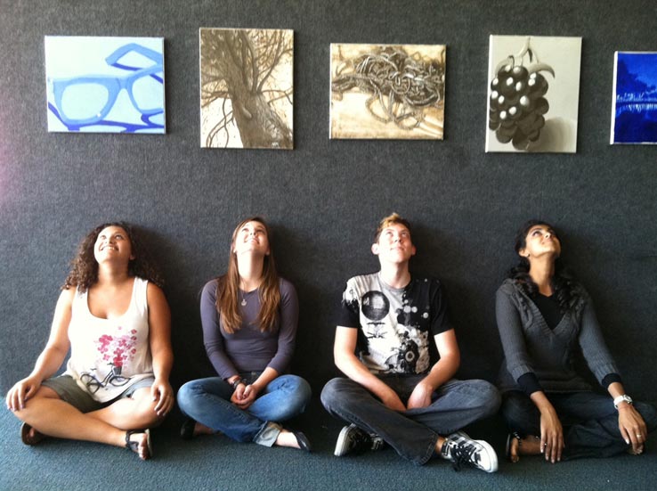 Students sitting beneath paintings displayed on a wall